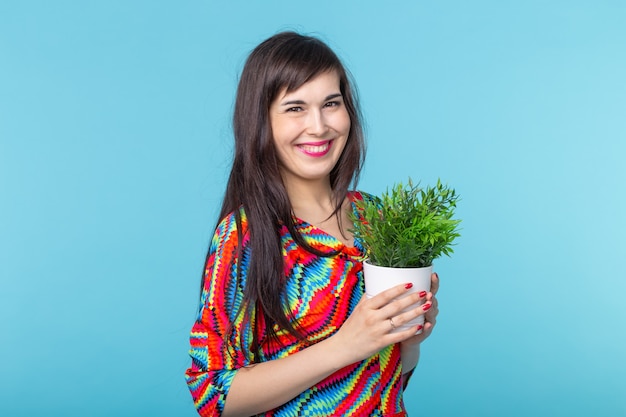 Une jeune femme joyeuse tient un pot avec une plante posant sur un fond bleu