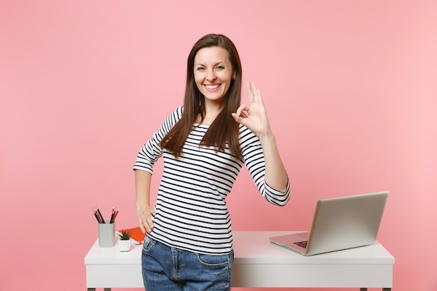 Jeune femme joyeuse en vêtements décontractés montrant un travail de signe OK, debout près d'un bureau blanc avec un ordinateur portable