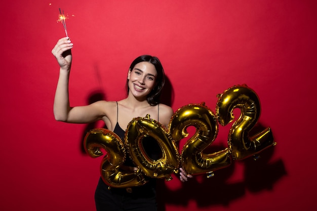 Une jeune femme joyeuse vêtue d'une robe noire tient les chiffres 2022 et des cierges magiques.