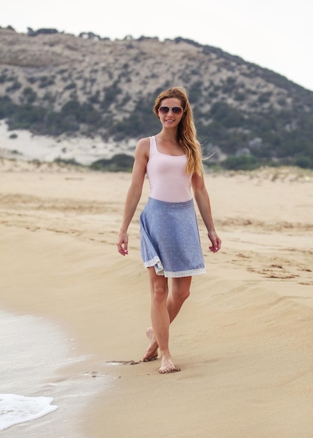 Jeune femme en jupe bleue, t-shirt et lunettes de soleil marchant sur la plage, souriante, temps couvert, colline derrière elle.