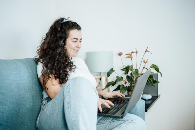 Jeune femme latina indépendante travaillant sur ordinateur tout en étant assise sur le canapé confortable à la maison sur des vêtements à la mode modernes. Concept de travail à distance à domicile, les jeunes travaillent en ligne