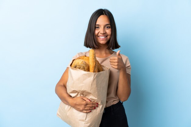 Jeune femme latine l'achat de pains isolés sur bleu donnant un geste de pouce en l'air