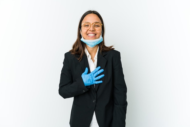 Jeune femme latine d'affaires portant un masque pour se protéger de covid isolé sur un mur blanc rit fort en gardant la main sur la poitrine.