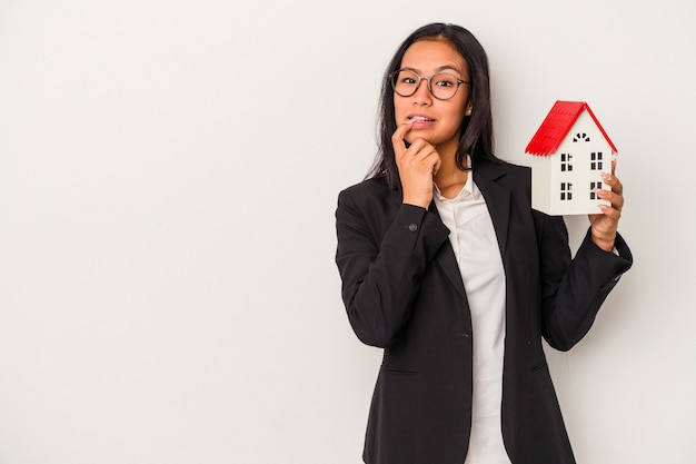 Jeune femme latine d'affaires tenant une maison de jouet isolée sur fond blanc détendue en pensant à quelque chose en regardant un espace de copie.