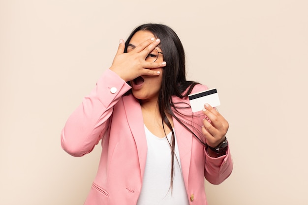 Jeune femme latine ayant l'air choquée, effrayée ou terrifiée, couvrant le visage avec la main et regardant entre les doigts