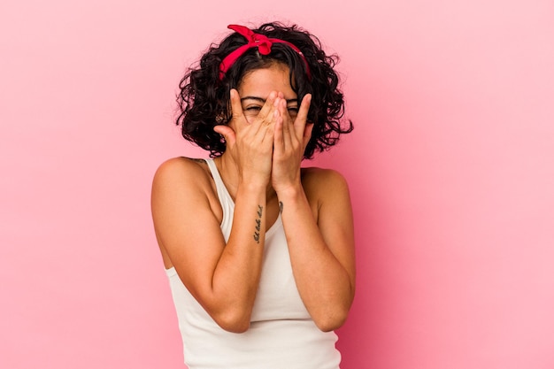 Jeune femme latine bouclée isolée sur fond rose clignote à la caméra à travers les doigts, visage couvrant embarrassé.