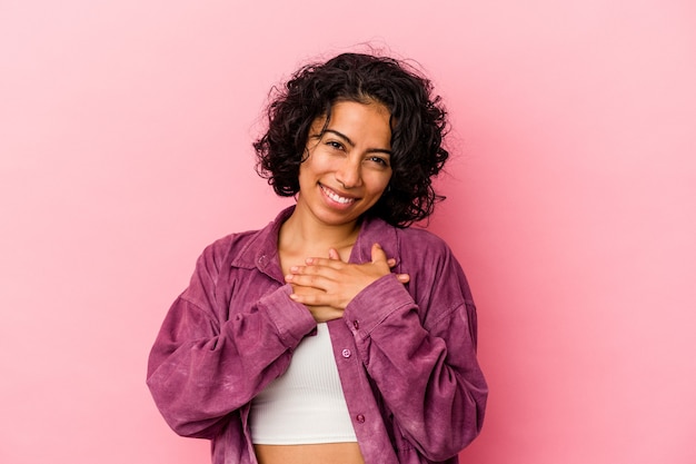 Jeune femme latine bouclée isolée sur fond rose a une expression amicale, appuyant la paume sur la poitrine. Notion d'amour.