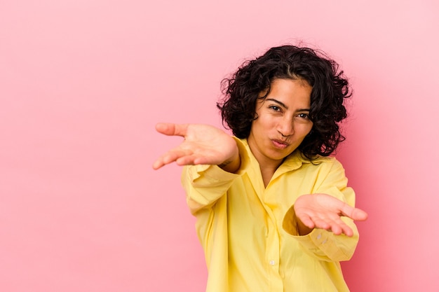 Jeune femme latine bouclée isolée sur fond rose pliant les lèvres et tenant les paumes pour envoyer un baiser aérien.