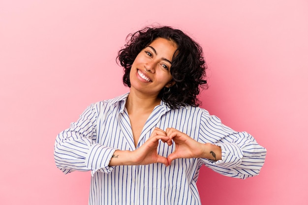 Jeune femme latine bouclée isolée sur fond rose souriant et montrant une forme de coeur avec les mains.