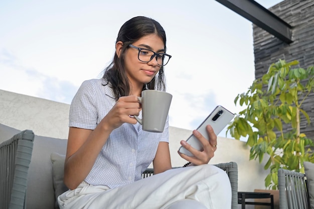 Jeune femme latine buvant du café tout en vérifiant les nouvelles sur son smartphone assis au coucher du soleil