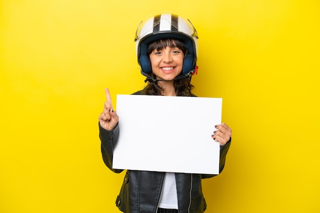 Jeune femme latine avec un casque de moto isolé sur fond jaune tenant une pancarte vide et pointant vers le haut