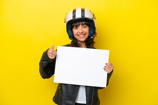 Jeune femme latine avec un casque de moto isolé sur fond jaune tenant une pancarte vide avec le pouce vers le haut