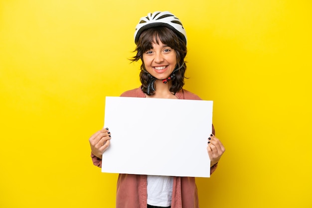 Jeune femme latine cycliste isolée sur fond jaune tenant une pancarte vide avec une expression heureuse