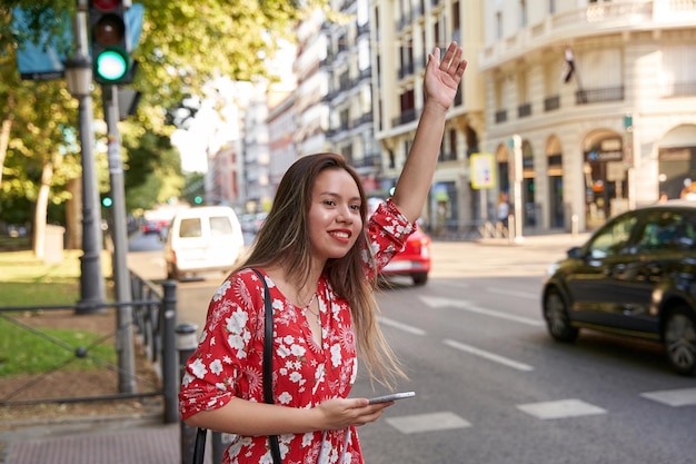 Jeune femme latine dans la rue et levant le bras pour attirer l'attention d'un taxi Elle tient un téléphone intelligent sur son autre main Concept d'application de taxi