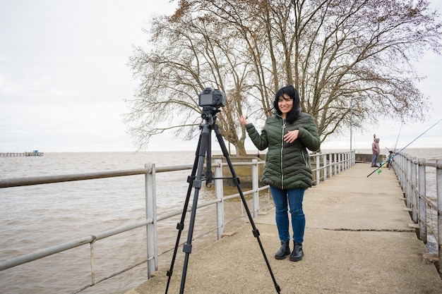 une jeune femme latine debout sur une jetée avec son appareil photo sur un trépied faisant un blog vidéo