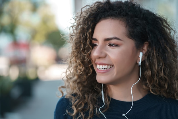 Jeune femme latine écoutant de la musique avec des écouteurs tout en se tenant à l'extérieur dans la rue