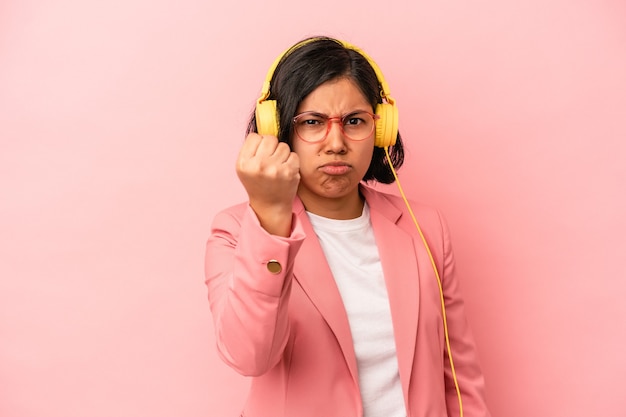 Jeune femme latine écoutant de la musique isolée sur fond rose montrant le poing à la caméra, expression faciale agressive.