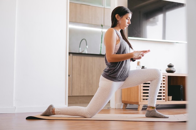 Jeune femme latine étirant ses jambes à la maison sur un tapis de yoga tout en discutant sur son téléphone