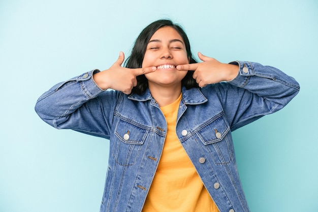 Jeune femme latine isolée sur fond bleu sourit, pointant du doigt la bouche.