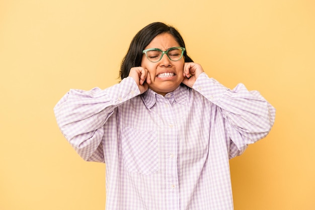 Jeune femme latine isolée sur fond jaune couvrant les oreilles avec les mains.