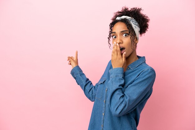 Jeune femme latine isolée sur fond rose avec une expression de surprise tout en pointant le côté