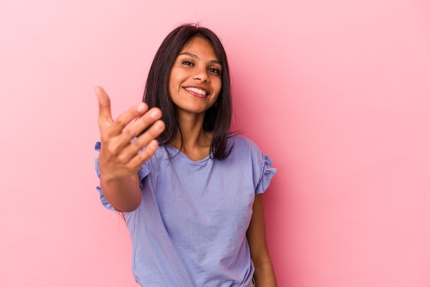 Jeune femme latine isolée sur fond rose s'étendant la main à la caméra en geste de salutation.