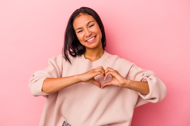 Jeune femme latine isolée sur fond rose souriante et montrant une forme de coeur avec les mains.