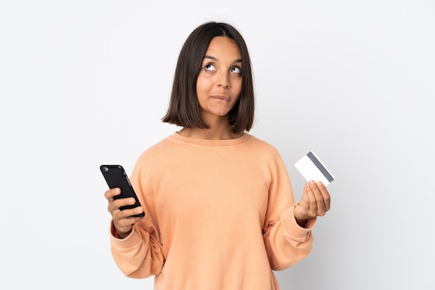 Jeune femme latine isolée sur un mur blanc acheter avec le mobile avec une carte de crédit tout en pensant