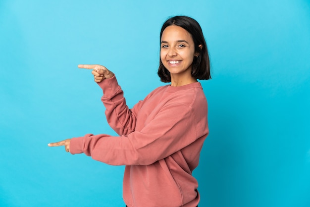 Jeune femme latine isolée sur le mur bleu pointant le doigt sur le côté et présentant un produit