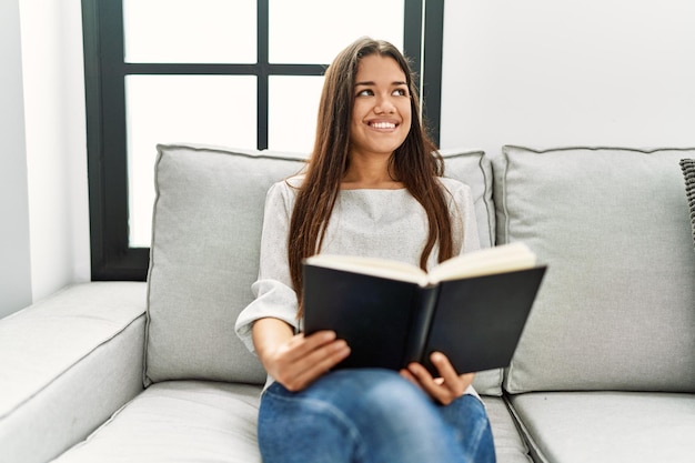 Jeune femme latine lisant un livre assis sur un canapé à la maison