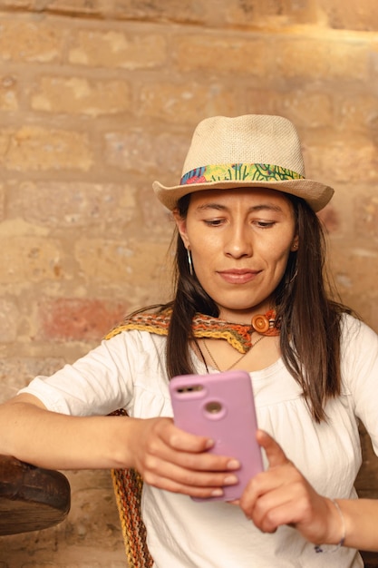 Jeune femme latine portant un chapeau assis sur une chaise en bois regardant son téléphone portable contre le mur de boue