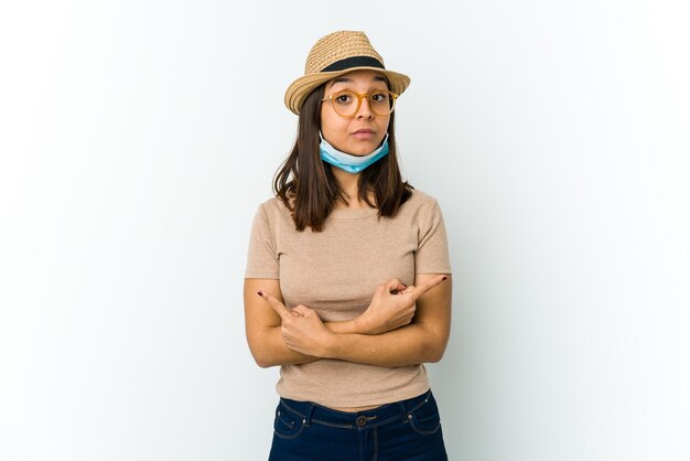 Jeune femme latine portant un chapeau et un masque pour se protéger de covid isolé sur fond blanc pointe sur le côté, essaie de choisir entre deux options.