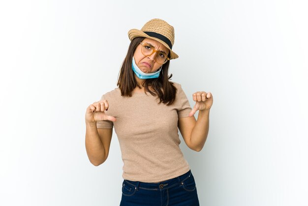 Jeune femme latine portant un chapeau et un masque pour se protéger de covid isolé sur un mur blanc montrant le pouce vers le bas et exprimant l'aversion.