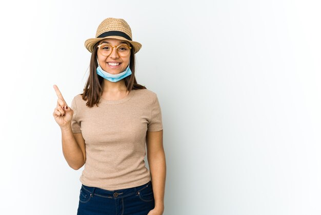 Jeune femme latine portant un chapeau et un masque pour se protéger du covid isolé sur fond blanc montrant le numéro un avec le doigt.