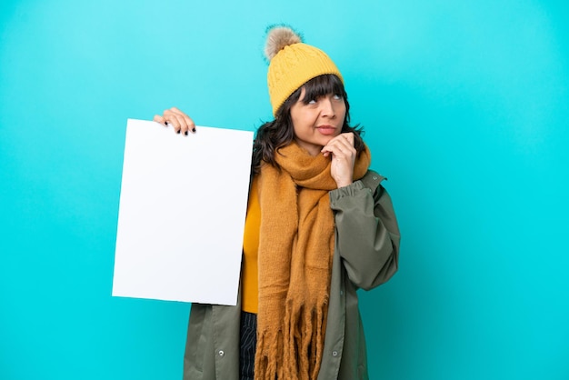 Photo jeune femme latine portant une veste d'hiver isolée sur fond bleu tenant une pancarte vide et pensant