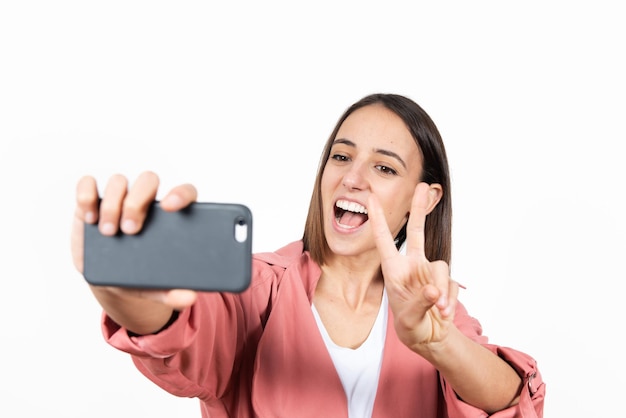 Jeune femme latine prenant un selfie tout en faisant le signe de la victoire sur fond blanc.