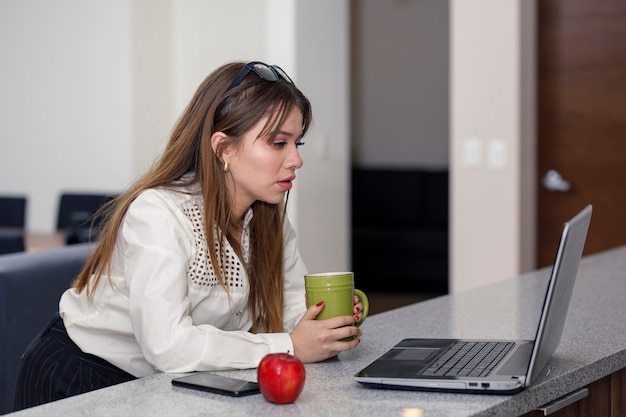 Jeune femme latine prenant une tasse de café tout en vérifiant son courrier sur son ordinateur portable depuis la cuisine