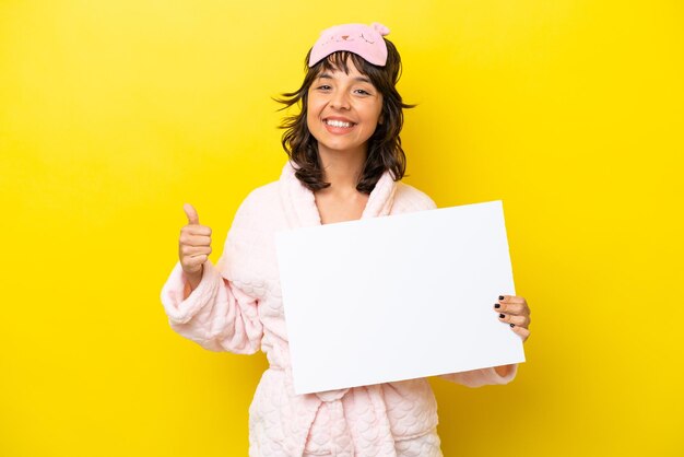 Jeune femme latine en pyjama isolé sur fond jaune tenant une pancarte vide avec le pouce vers le haut