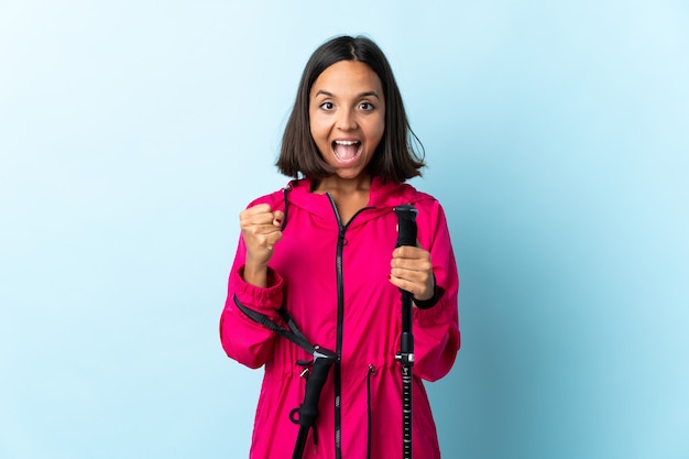 Jeune femme latine avec sac à dos et bâtons de randonnée bleu célébrant une victoire en position de vainqueur