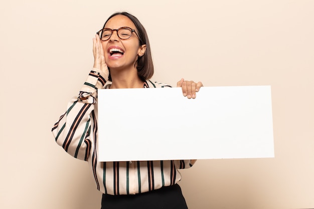 Jeune femme latine se sentant heureuse, excitée et positive, donnant un grand cri avec les mains à côté de la bouche, appelant