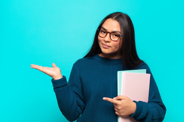 Jeune femme latine souriant joyeusement et pointant pour copier l'espace sur la paume sur le côté, montrant ou faisant la publicité d'un objet