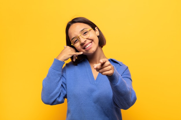 Jeune femme latine souriant joyeusement et pointant vers la caméra tout en faisant un appel, vous geste plus tard, parler au téléphone