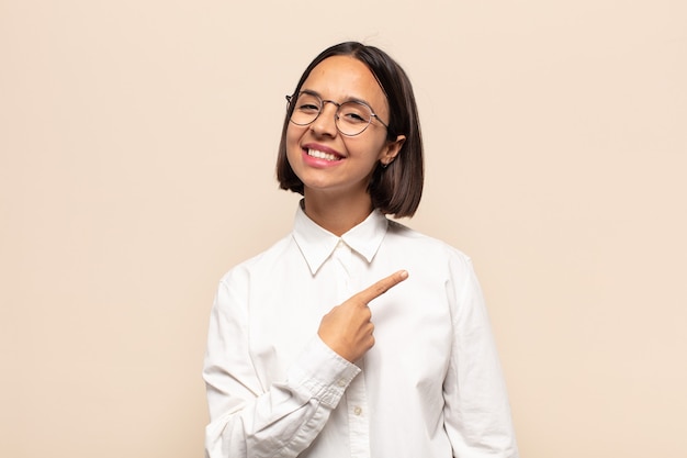 Jeune femme latine souriant joyeusement, se sentant heureux et pointant vers le côté et vers le haut, montrant l'objet dans l'espace de copie