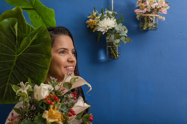 Jeune femme latine souriante près de feuilles tropicales vertes et fleurs visage fermé sur fond bleu