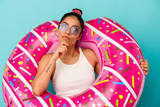 Jeune femme latine tenant un beignet gonflable isolé sur fond bleu regardant de côté avec une expression douteuse et sceptique.