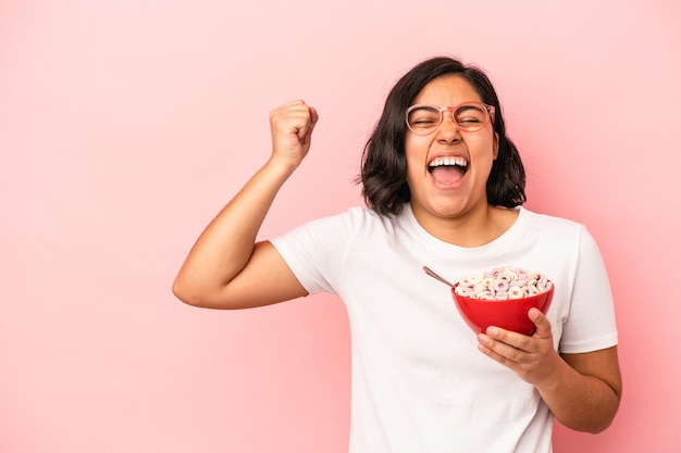 Jeune femme latine tenant des céréales isolées sur fond rose levant le poing après une victoire, concept gagnant.