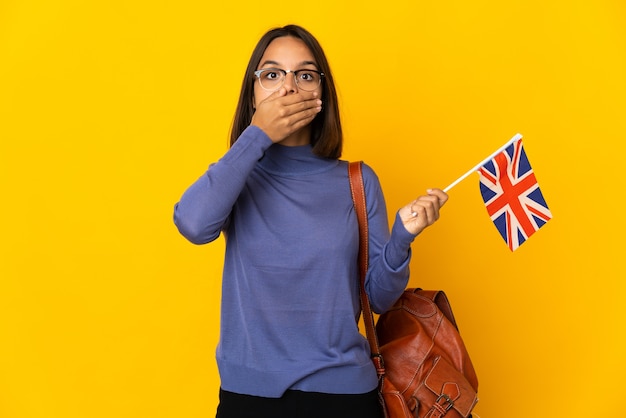 Jeune femme latine tenant un drapeau du Royaume-Uni isolé sur fond jaune couvrant la bouche avec la main