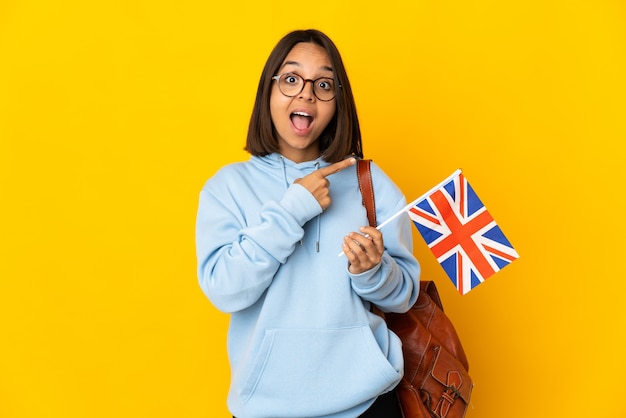 Jeune femme latine tenant un drapeau du Royaume-Uni isolé sur fond jaune surpris et côté pointant