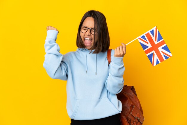 Jeune femme latine tenant un drapeau du Royaume-Uni isolé sur un mur jaune célébrant une victoire