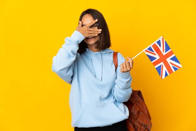 Jeune femme latine tenant un drapeau du Royaume-Uni isolé sur un mur jaune couvrant les yeux par les mains et souriant